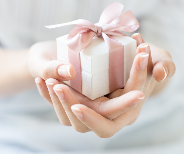 The image is focused on two cupped hands holding a small white gift box with a pale pink ribbon wrapped around it