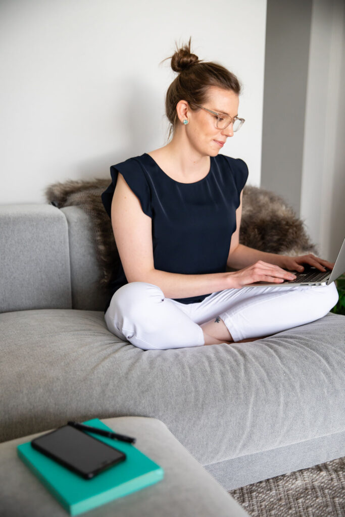 A woman sitting crossed-legged on a grey couch looking at a laptop whilst she writes. She is wearing white jeans and a navy blue blouse. There is a furry blanket behind her.