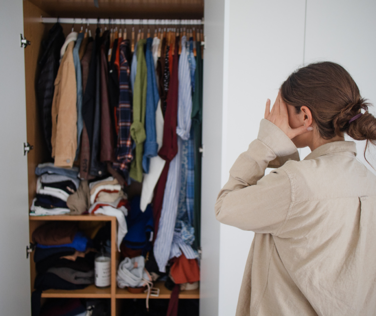 A woman is a light brown is standing facing an open closet. She is holding her hands to her face. The closet in front of her is messy with clothing haphazardly placed inside.